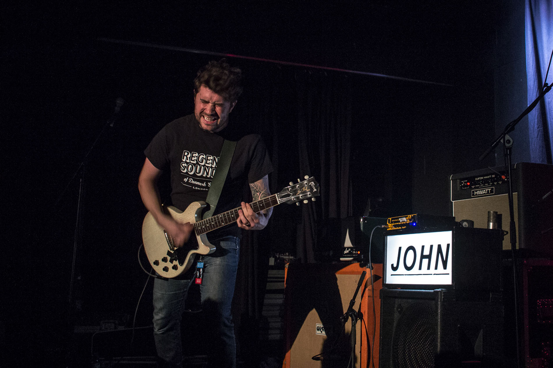 Guitarist from JOHN performing at Hard Club beside an illuminated JOHN sign.